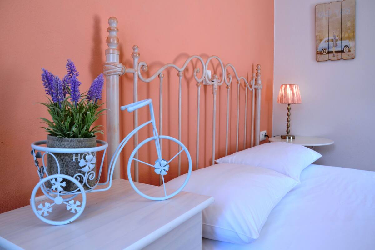 Een slaapkamer met een bed, een nachtkastje met een lamp, een decoratieve metalen fiets en een bloempot met plant.
