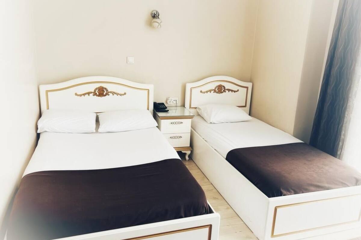 A hotel room with two single beds, each with a white headboard and a dark-colored bedspread. There is a nightstand with a lamp and a phone between the beds. The room has a plain wall and a window with a curtain.