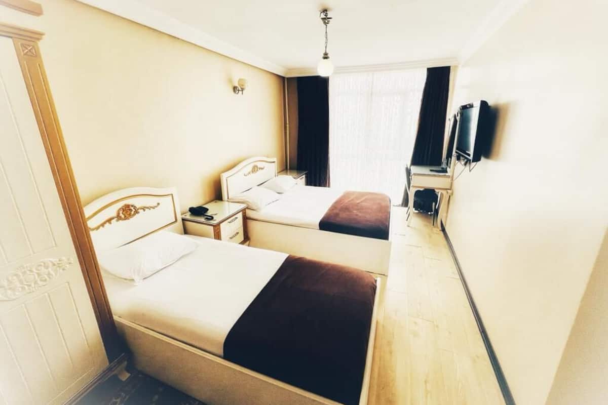 A hotel room with two beds, a bedside table, a nightstand, a TV mounted on the wall, and a window with curtains.