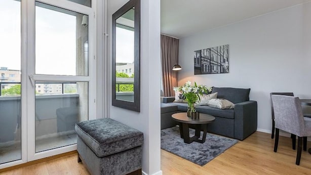 Una sala de estar moderna con un sofá gris, una mesa redonda y un reposapiés gris. Se ve un ventanal con vistas a edificios.