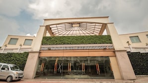A hotel entrance with a glass facade, a covered entrance area, and a green hedge. - Hotel Celebration Gardens (New Delhi)