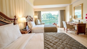 A hotel room with two beds, a desk, a chair, a lamp, and a window with a view of greenery. - Hotel Celebration Gardens (New Delhi)