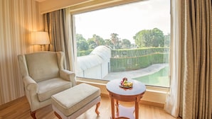 A room with a large window, a chair, an ottoman, and a table with fruit. - Hotel Celebration Gardens (New Delhi)