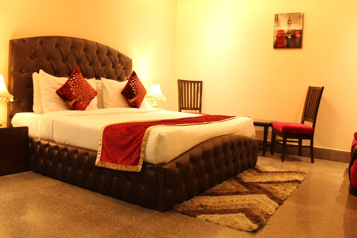 A neatly made bed with a red throw blanket, a wooden nightstand with a lamp, a chair, and a small table with a red cushion.