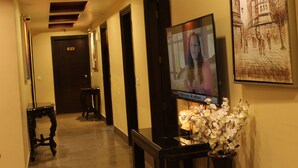 A hotel corridor with a television displaying a news program, a clock on the wall, and a framed picture of a building. - Karan Holidays (New Delhi)