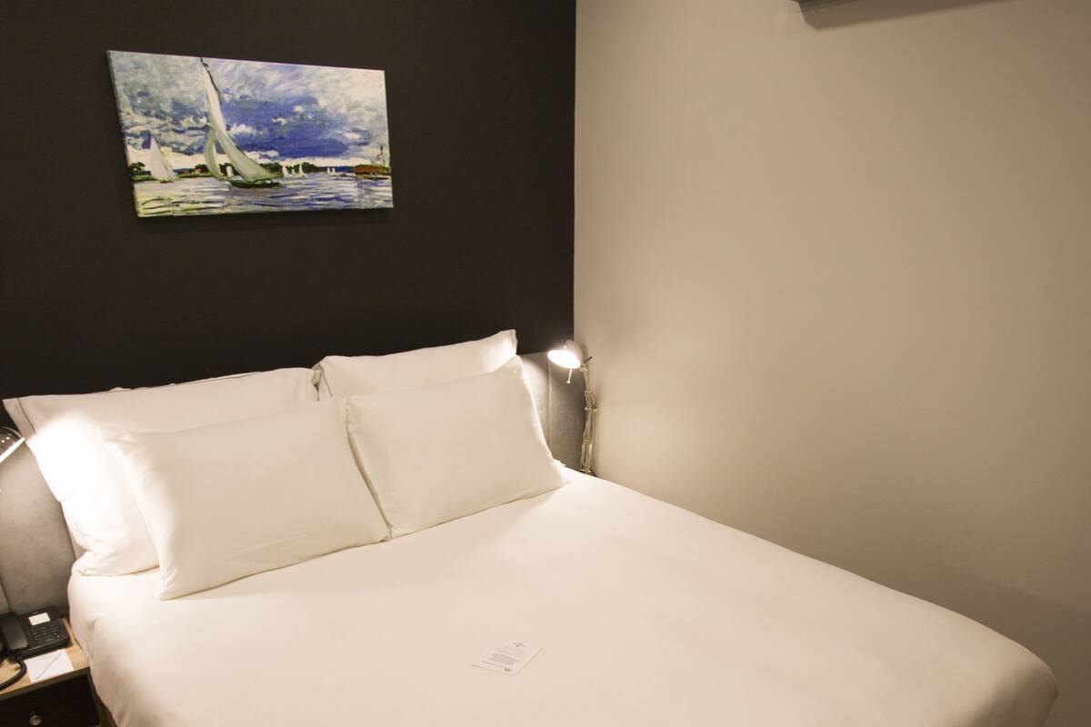 A neatly made bed in a hotel room with a painting on the wall, a bedside lamp, and an air conditioning unit.