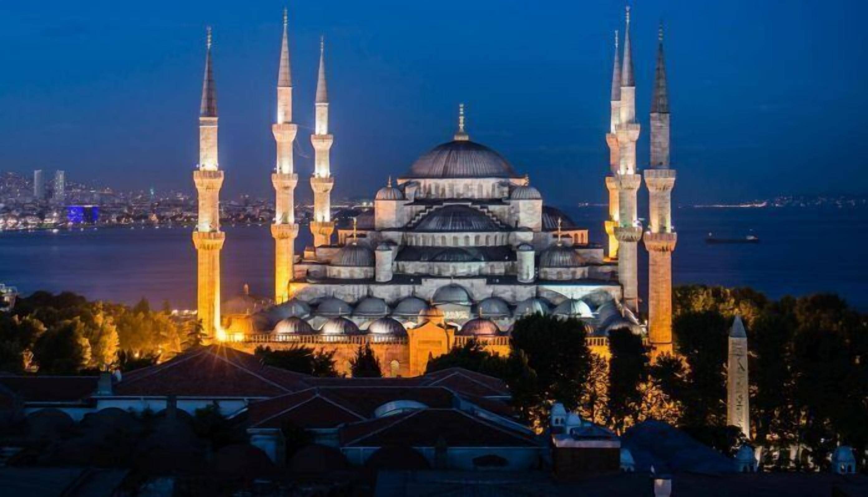 A grand mosque with multiple minarets and a large central dome, surrounded by water and urban skyline.