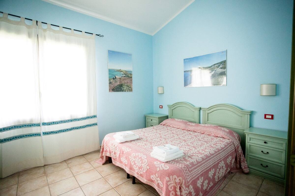 A bedroom with a bed, two nightstands, a window with curtains, and two wall-mounted lamps.
