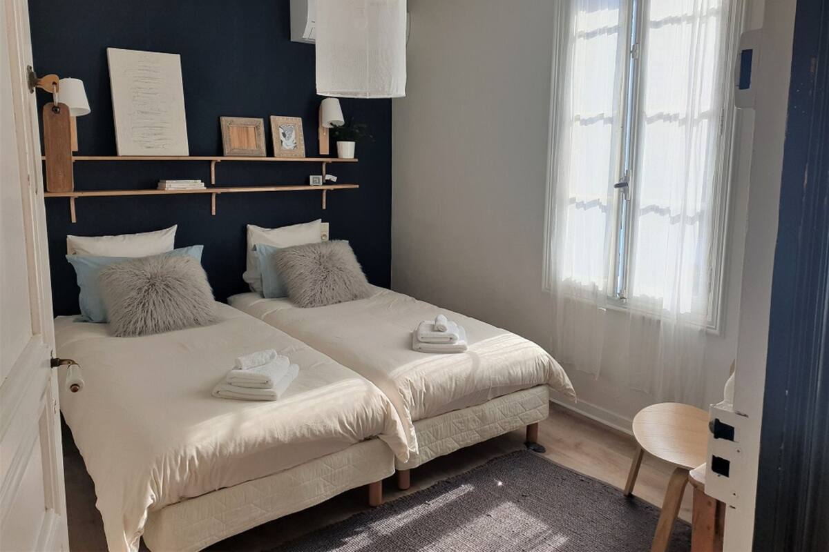 A bedroom with two beds, a bedside table, a chair, a window with sheer curtains, and wall-mounted shelves with decorative items.