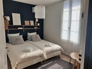A bedroom with two beds, a bedside table, a chair, a window with sheer curtains, and wall-mounted shelves with decorative items. - L'Abri Sous Roche - Chambres d'hôtes (Tautavel)