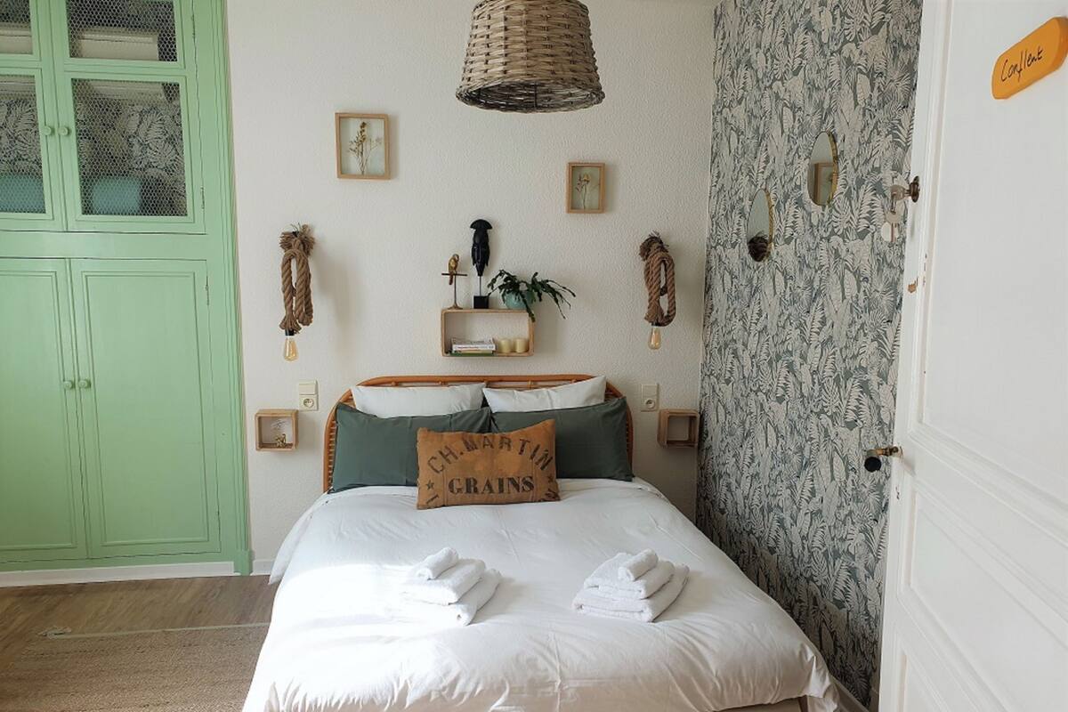 A neatly made bed with white linens and a pillow with 'CH. MARTIN' text, a green wardrobe, a wicker lamp, and a patterned wallpaper on the wall.
