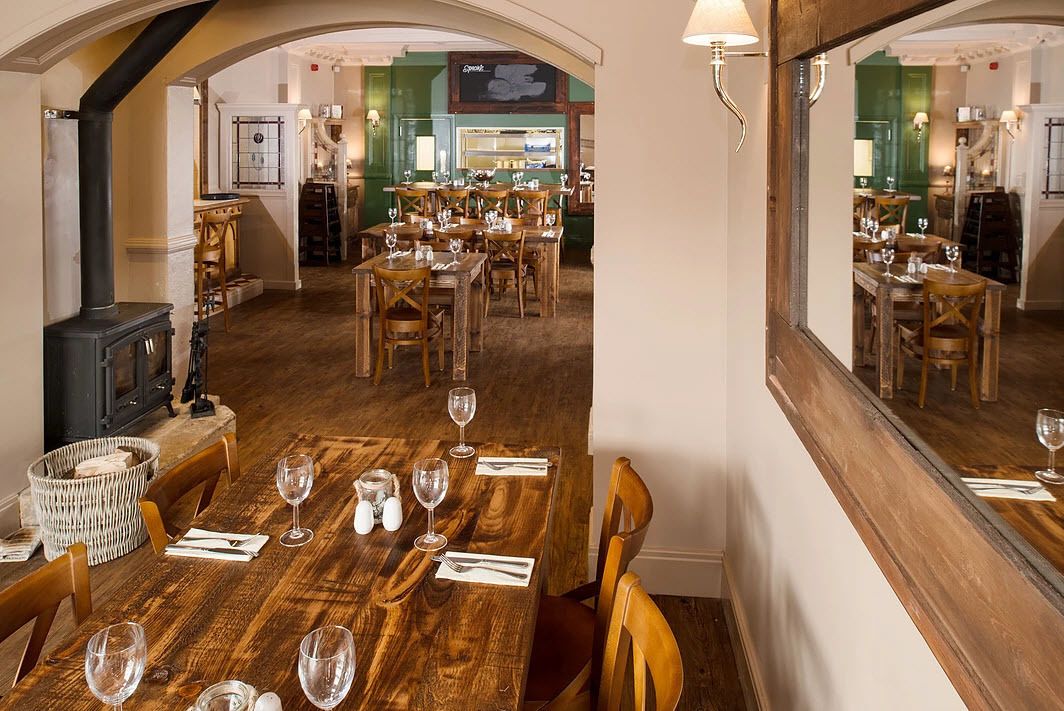 A restaurant with wooden tables and chairs, a fireplace, and a view of the kitchen in the background.
