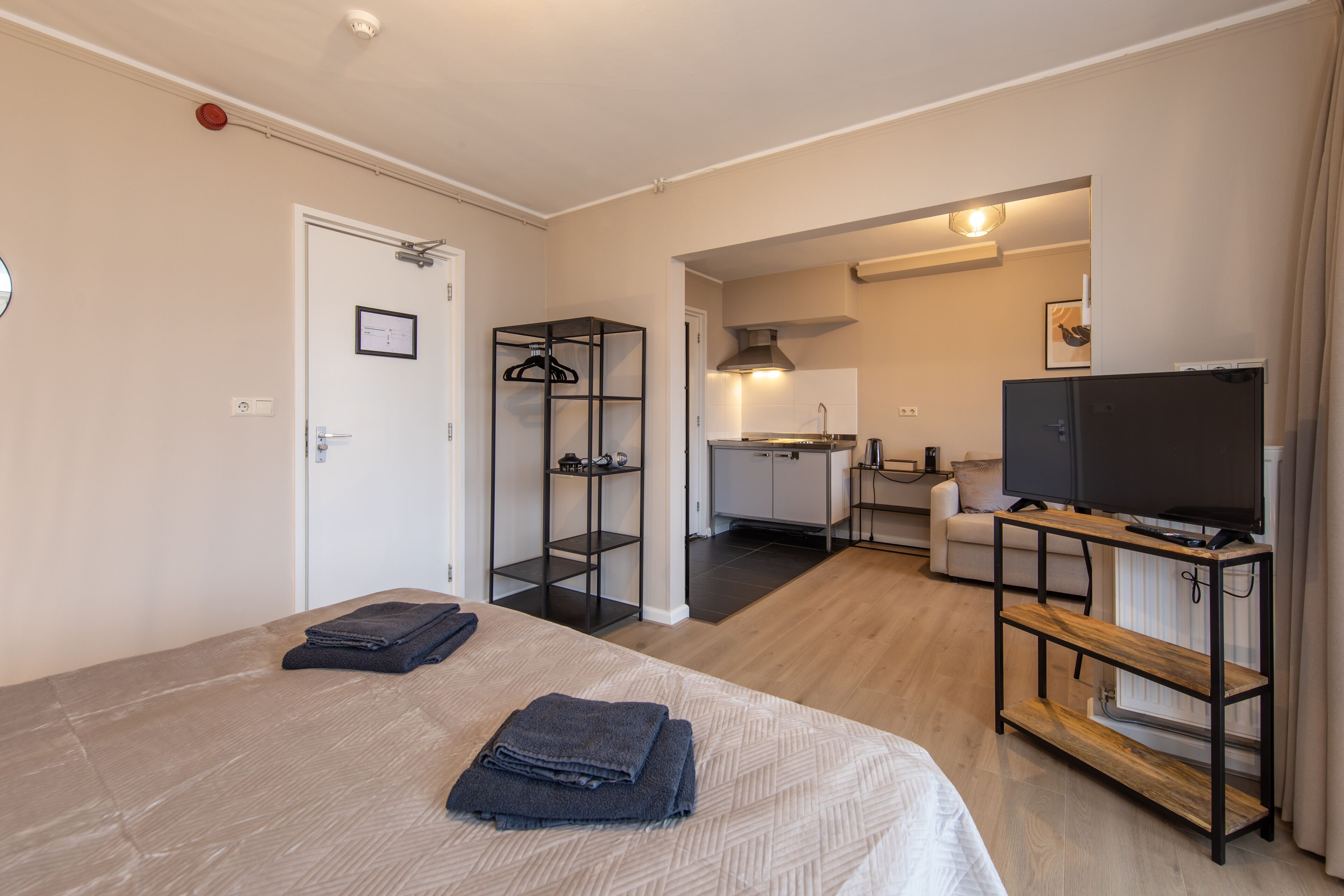 A modern hotel room with a large bed, a TV on a wooden stand, a kitchenette, and a separate living area with a sofa.