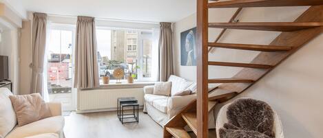 A living room with a sofa, a chair, a small table, a window with curtains, and a wooden staircase.