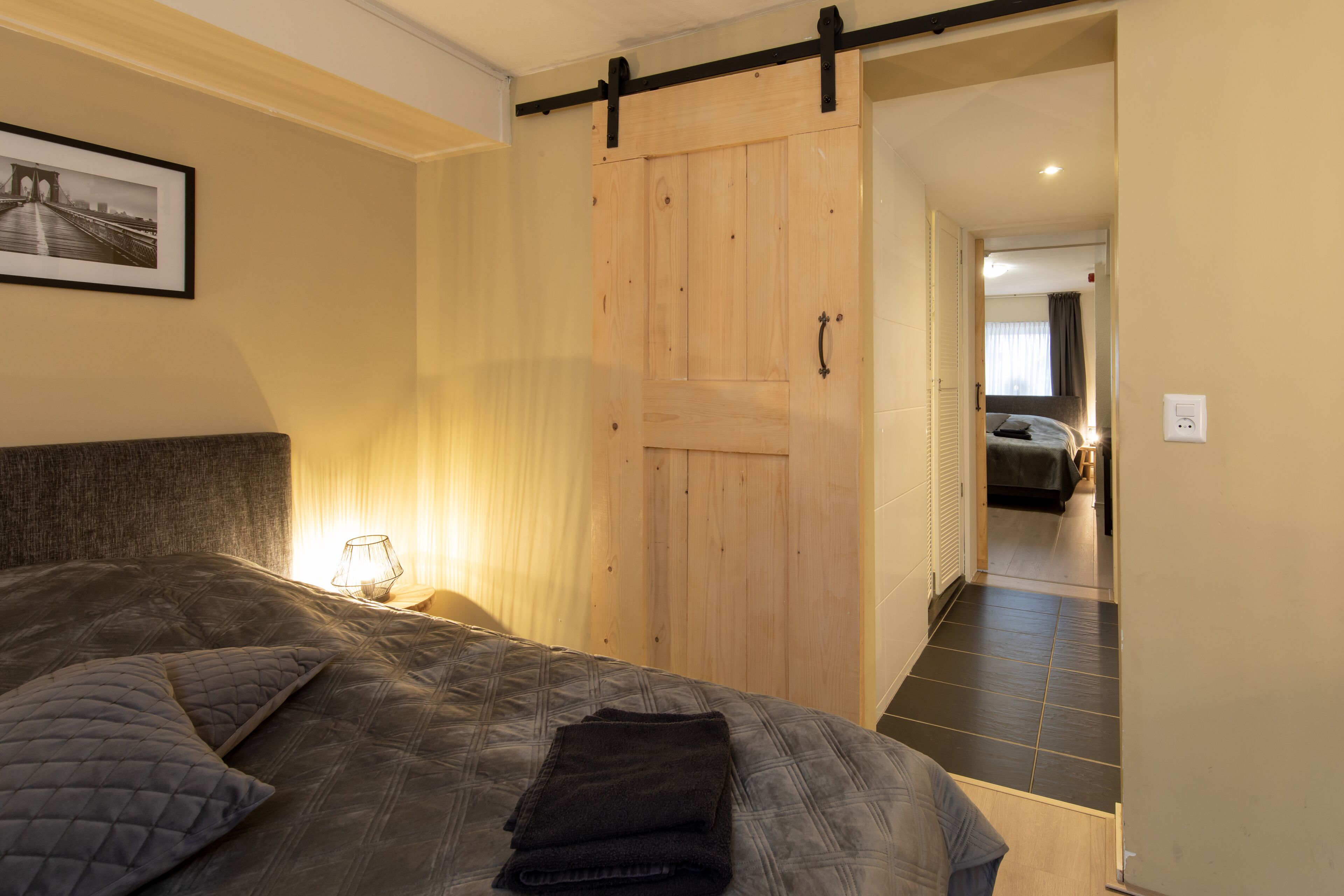 Een slaapkamer met een groot bed, een houten schuifdeur, een lijst met een afbeelding aan de muur, en een nachtkastje met een lamp.