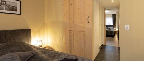 Een slaapkamer met een groot bed, een houten schuifdeur, een lijst met een afbeelding aan de muur, en een nachtkastje met een lamp.