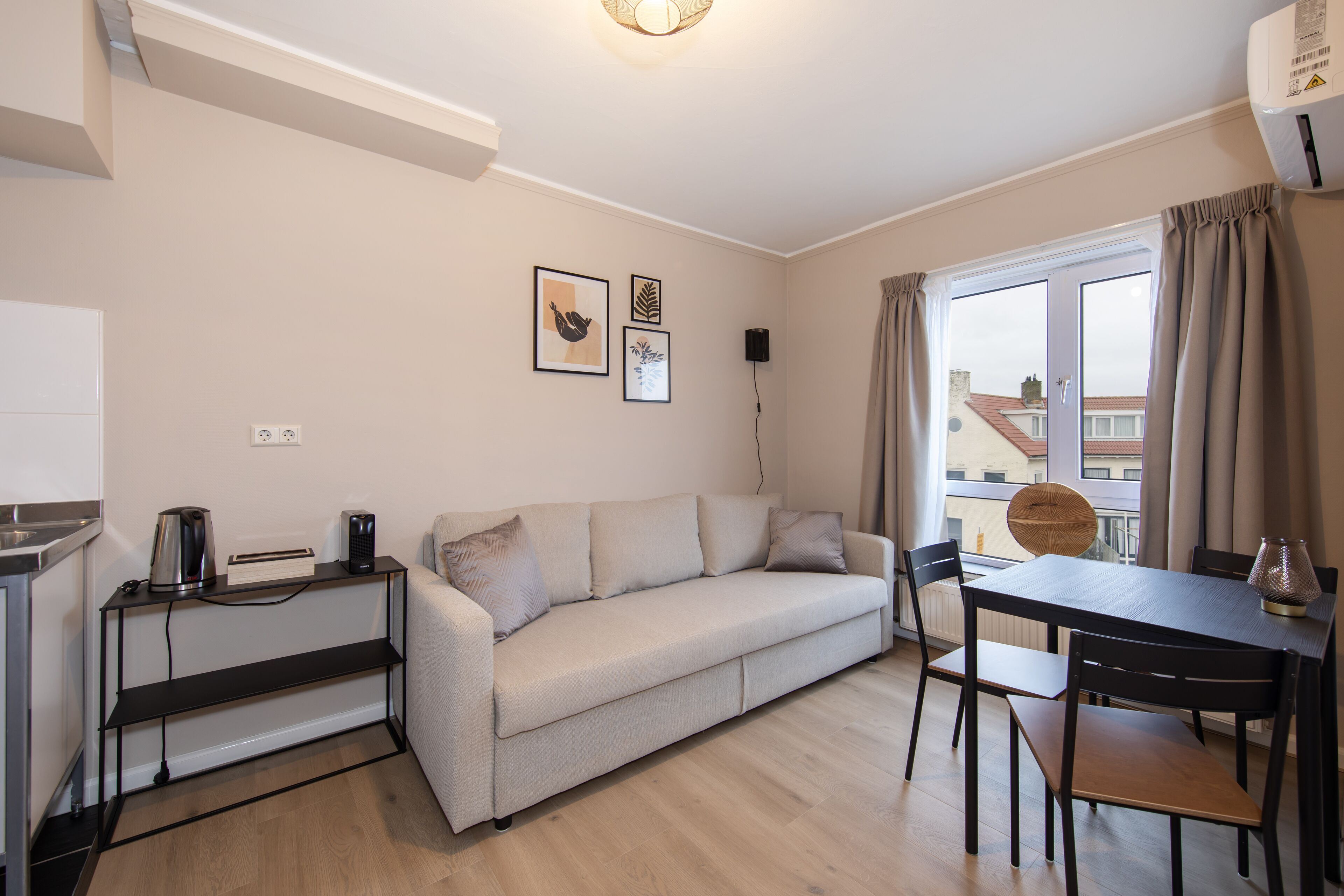 A modern living room with a sofa, a dining table, and chairs. There is a kitchen area with a kettle and a speaker on the wall.