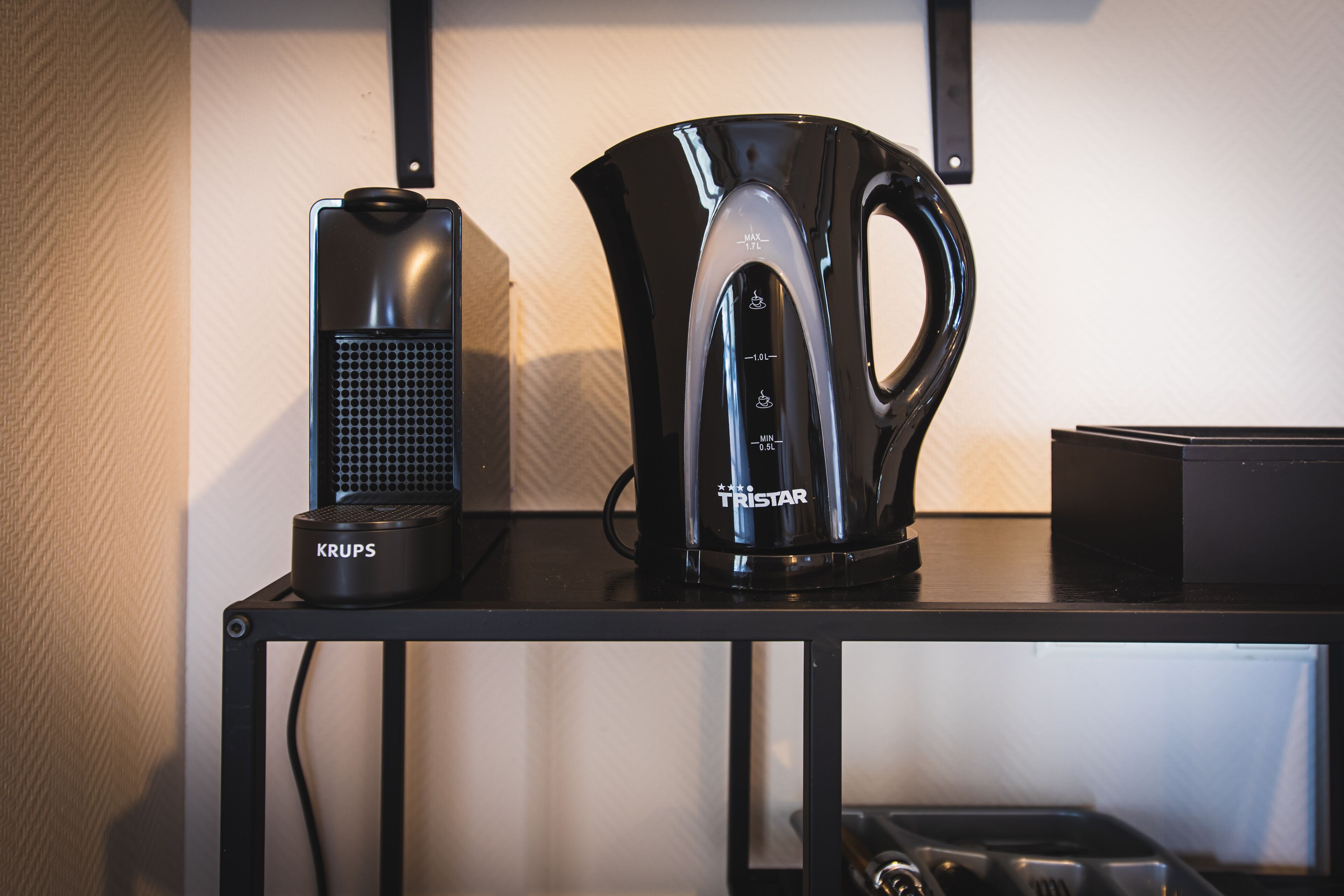 Eine Kaffeemaschine und ein Wasserkocher, beide schwarz, stehen auf einem schwarzen Metallregal.