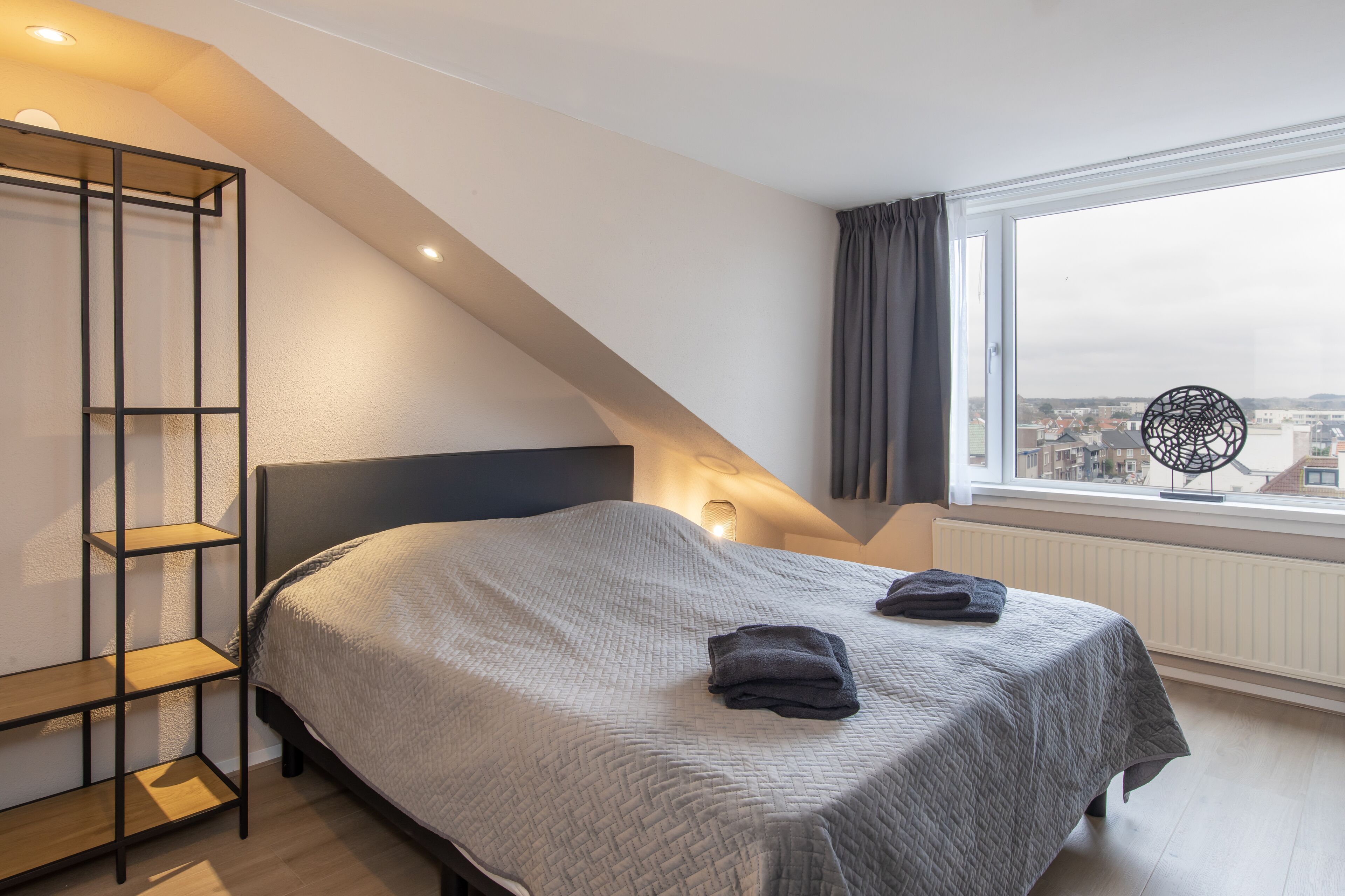 A bedroom with a bed, a wooden shelf unit, a window with a view of buildings, and a ceiling light.