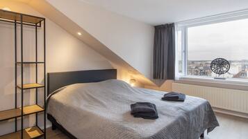A bedroom with a bed, a wooden shelf unit, a window with a view of buildings, and a ceiling light.