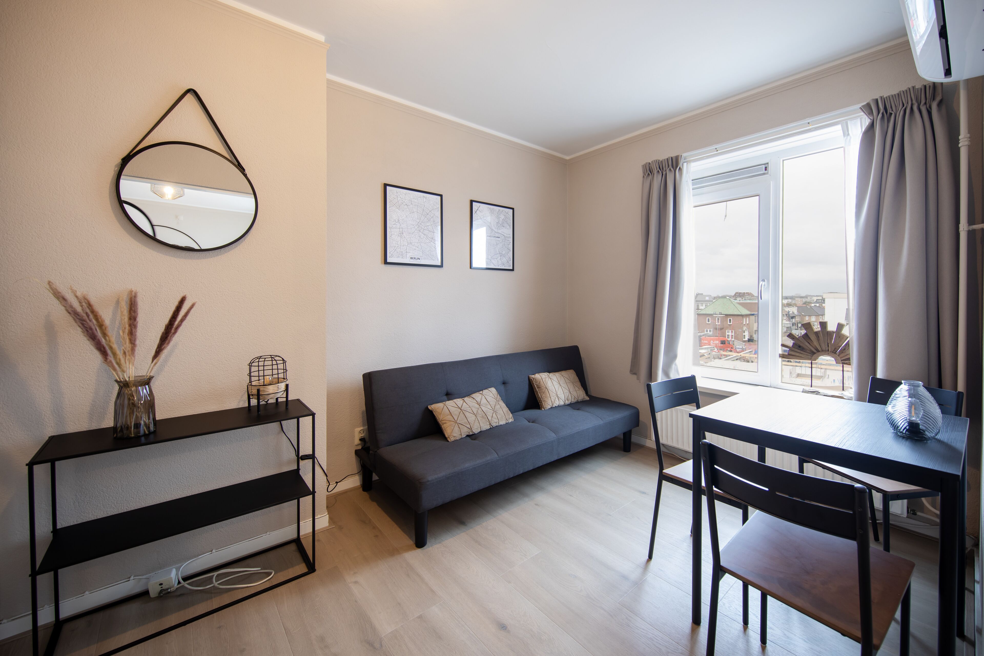 Ein modernes Wohnzimmer mit einer Couch, einem Esstisch und Stühlen. Ein TV-Schrank mit einer Vase und einer Lampe steht dort. An der Wand hängen zwei gerahmte Bilder, und ein Fenster mit Vorhängen ist zu sehen.
