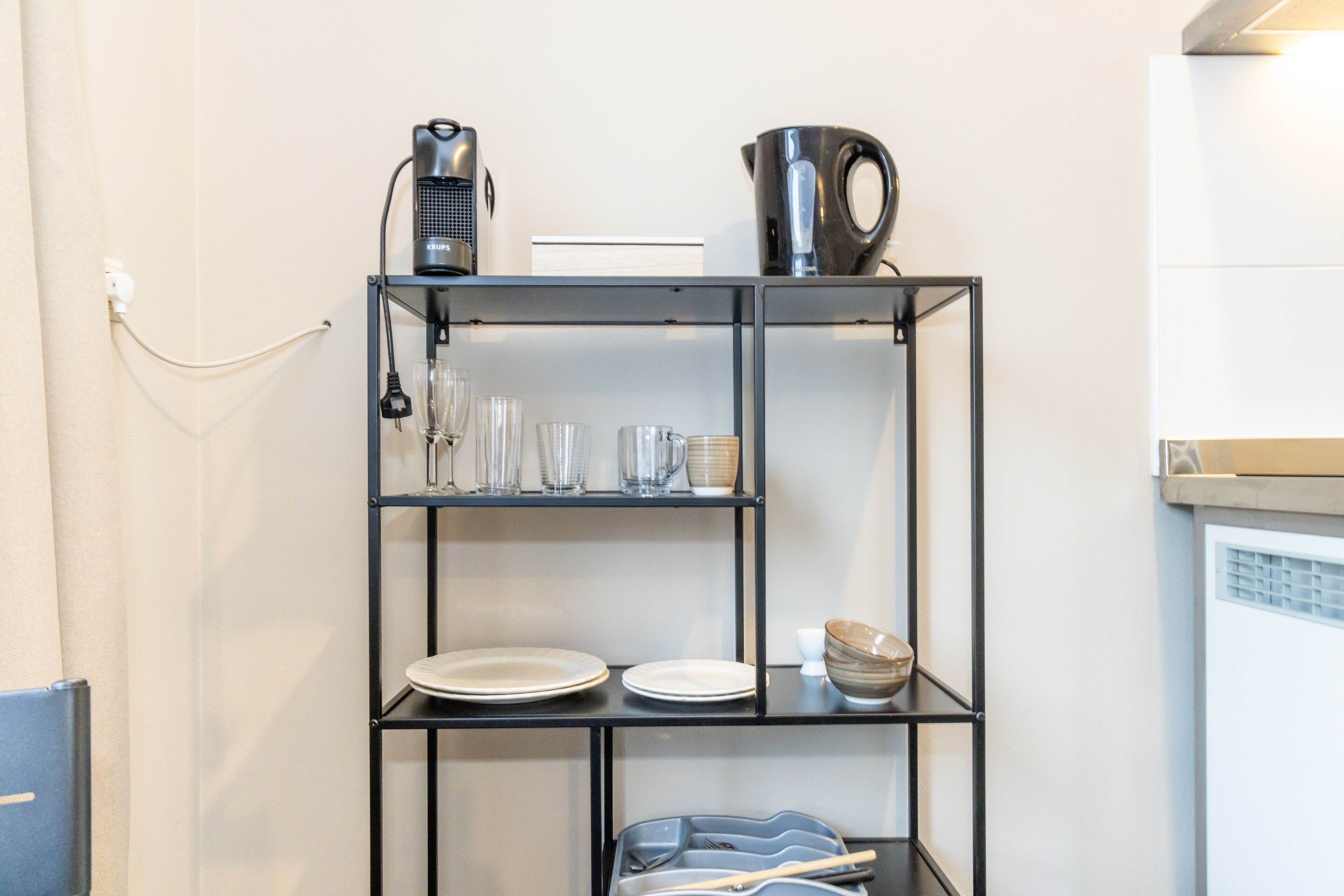 Ein schwarzes Metallregal mit verschiedenen Küchenutensilien, darunter Teller, Gläser, ein Wasserkocher und eine Kaffeemaschine.