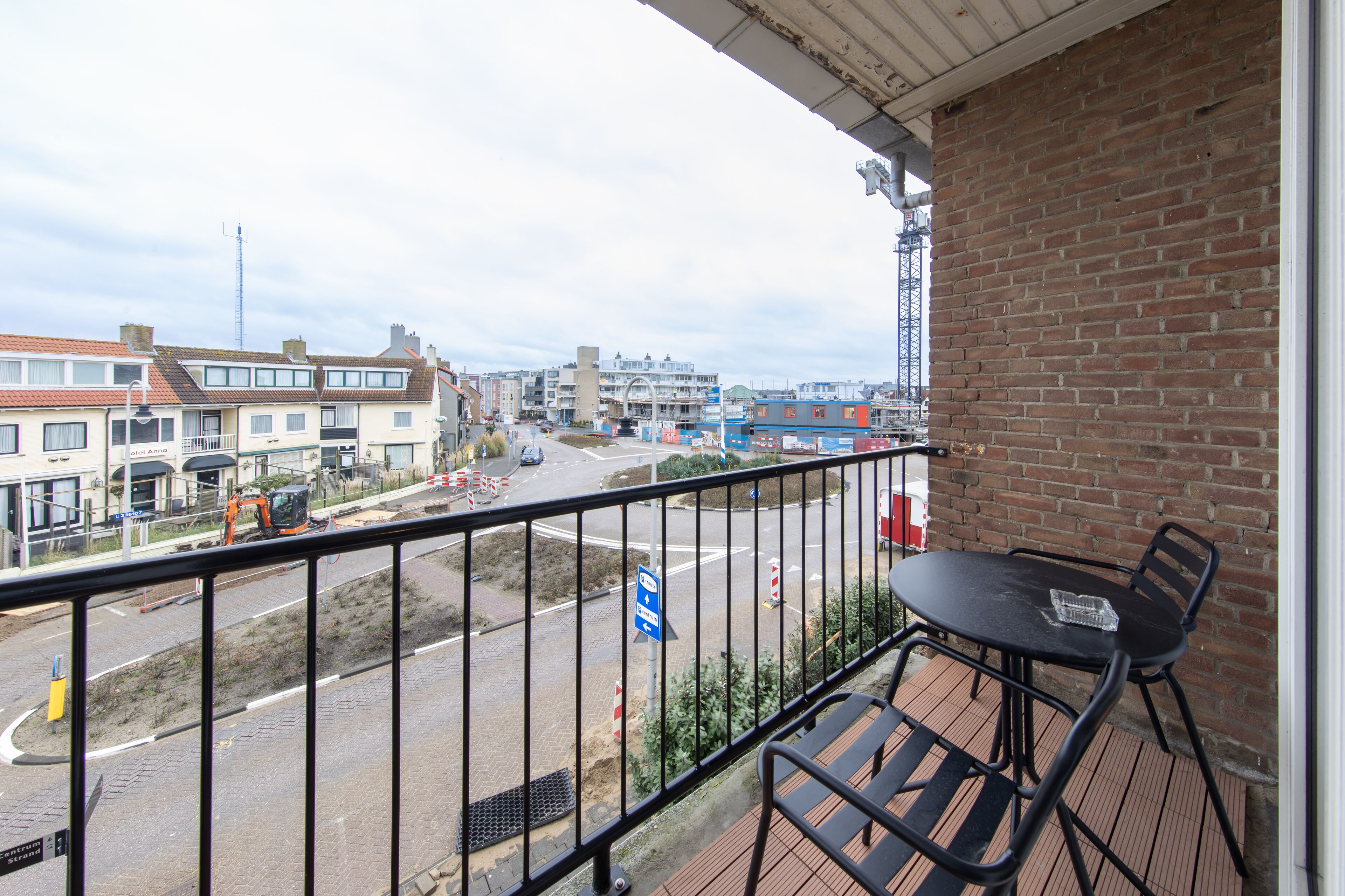 Un balcón con una mesa y sillas, con vista a una calle con edificios y obras en construcción.