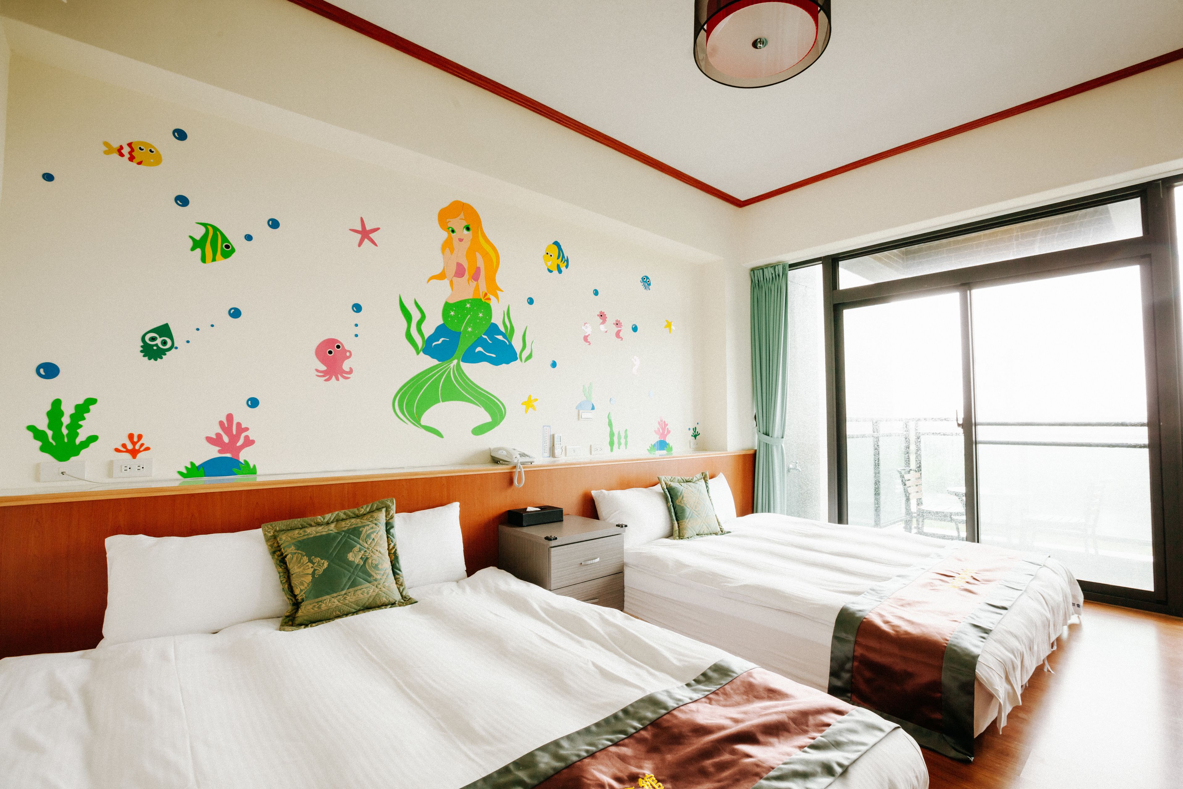 A hotel room with two beds, a wooden headboard, a wall mural of a mermaid, and a large window with a view.