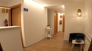 A hotel reception area with a reception desk, a sign indicating the reception, a small table with a bowl, a black chair, a plant on a stool, and a fire extinguisher. - Hostal Mónaco (Valladolid)