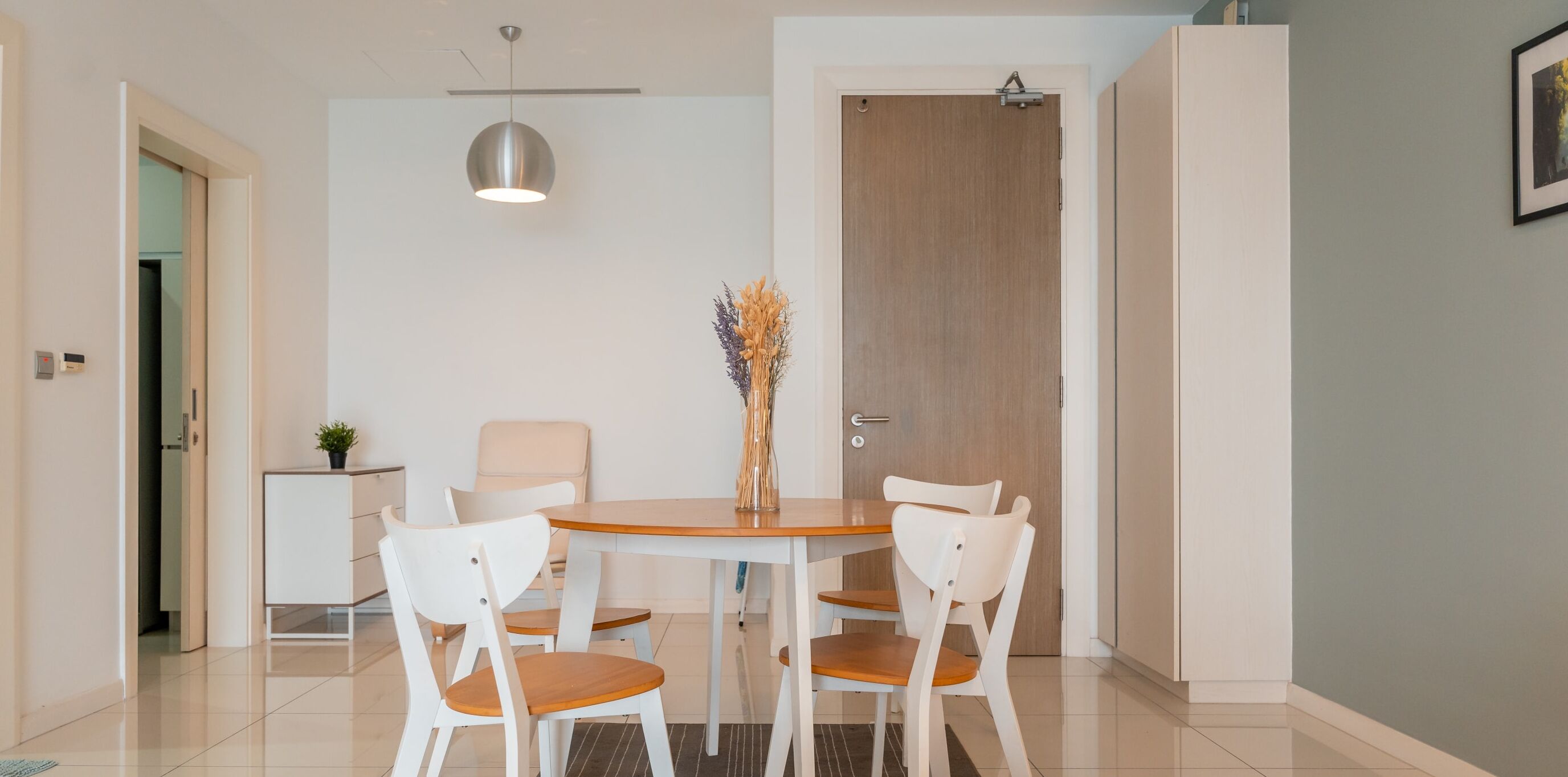 Un coin repas avec une table ronde, quatre chaises blanches, une petite table d’appoint avec une plante, un luminaire suspendu et une porte en bois.
