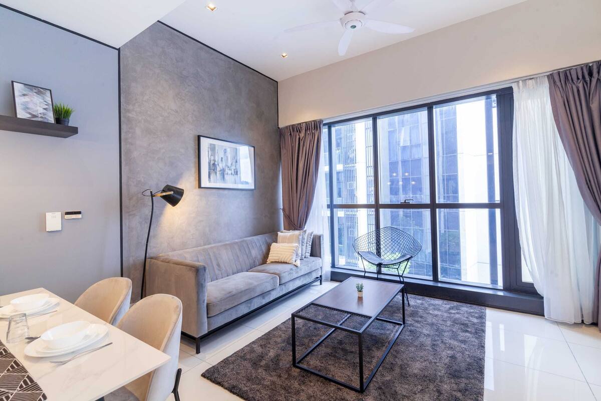 A modern living room with a grey sofa, a dining table set with plates, a coffee table, and a large window with curtains.