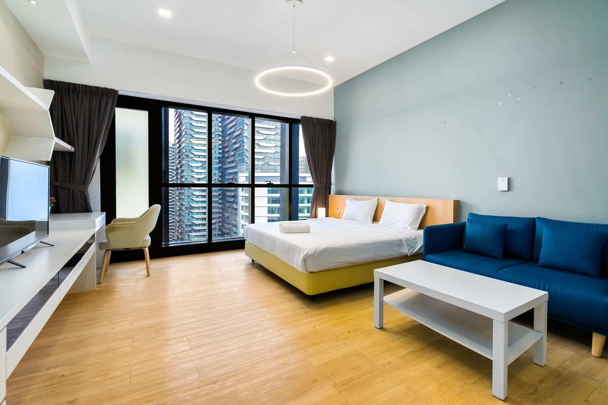 A modern hotel room with a bed, a blue sofa, a white coffee table, a desk with a TV, and large windows offering a city view.