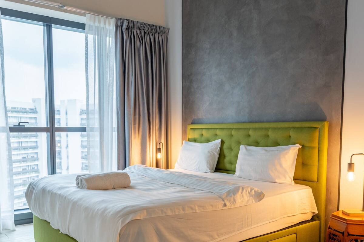 A hotel room with a large bed, a green upholstered headboard, white bedding, and a view of the city through large windows.