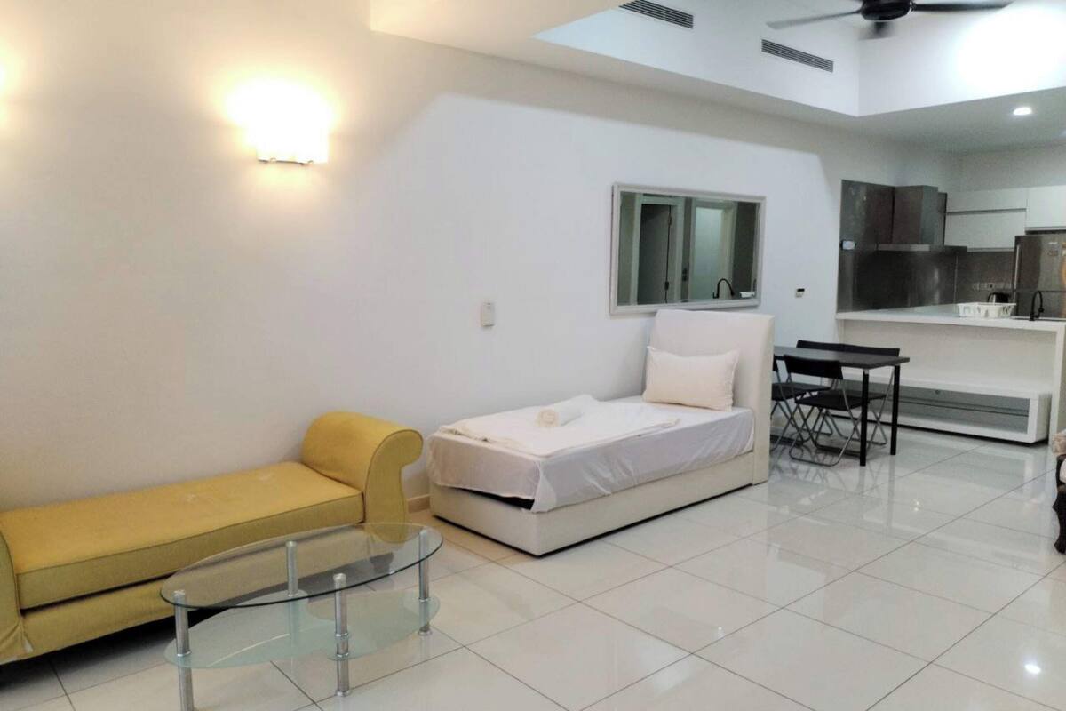 A modern, well-lit room with a bed, a sofa, a glass table, and a kitchenette area.