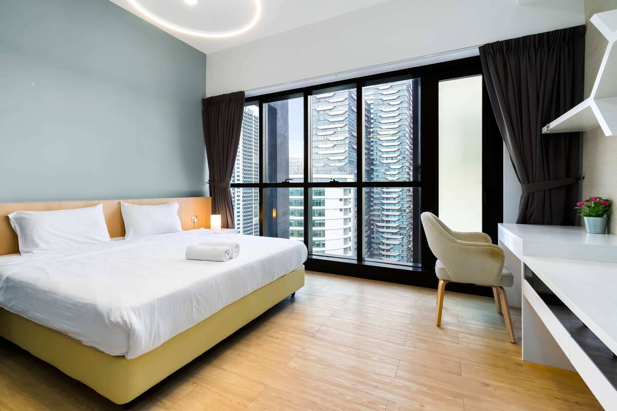 A modern hotel room with a large bed, a desk, and a view of tall buildings through large windows.