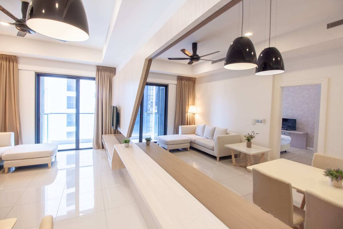 A modern hotel room with a sofa, a dining table, a TV, and a balcony with curtains.