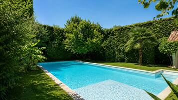 Una piscina rectangular con superficie de agua azul cristalina, rodeada de exuberantes árboles y césped.