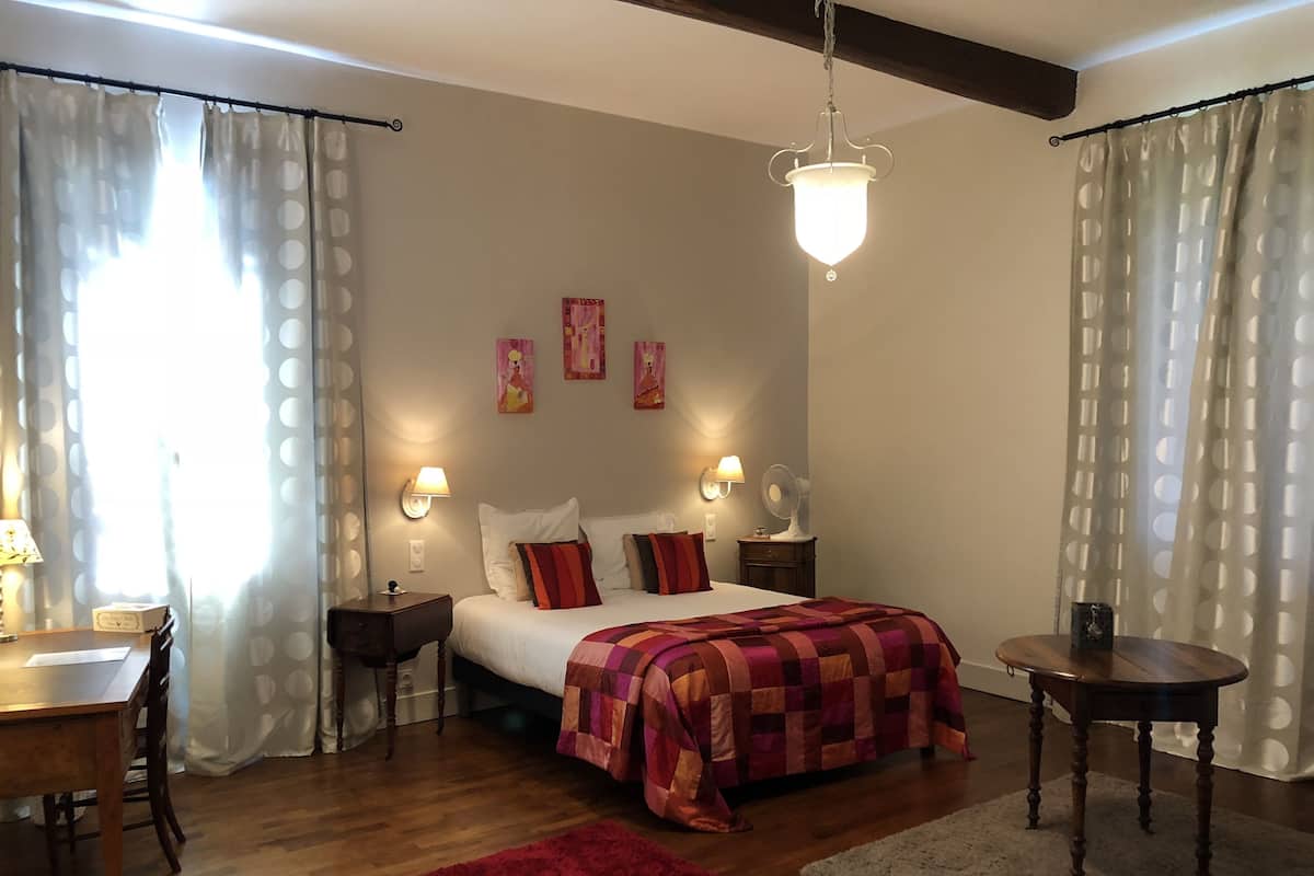 A bedroom with a bed, two bedside tables, a desk, a chair, a red rug, a hanging light fixture, and wall art.