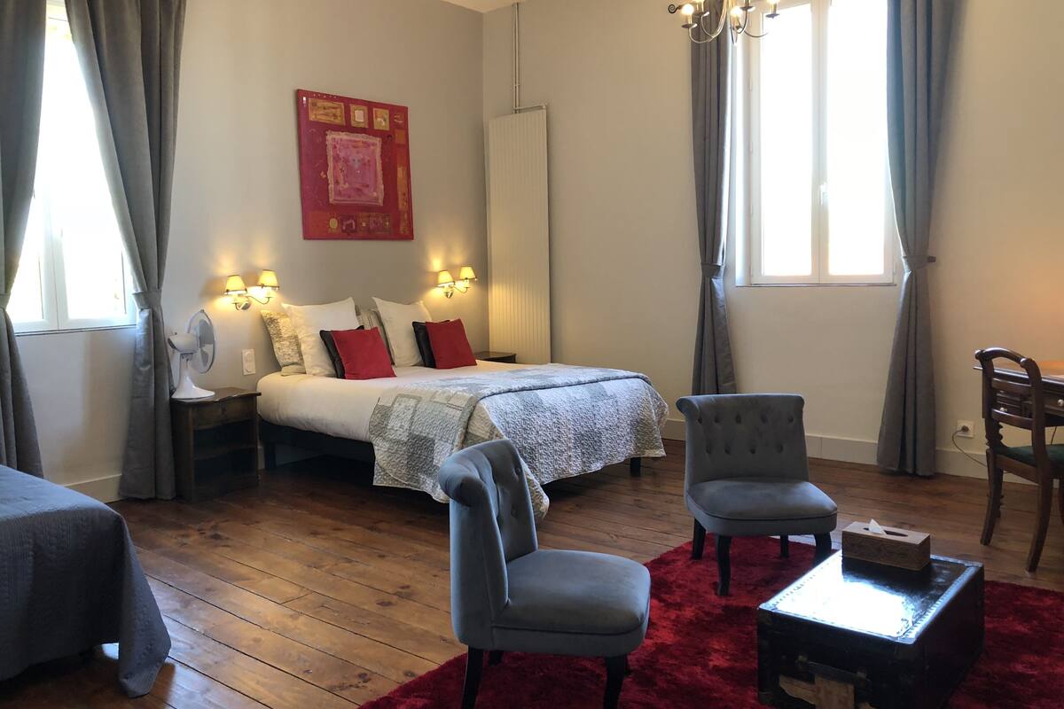 A bedroom with a bed, two bedside tables, a nightstand, a chair, a small table, a red rug, and a window with curtains.