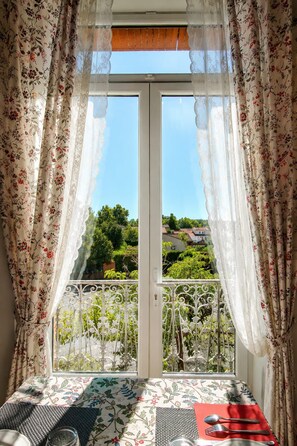 Un balcón con una mesa lista para comer, cortinas con estampado floral y vistas a zonas verdes y edificios.