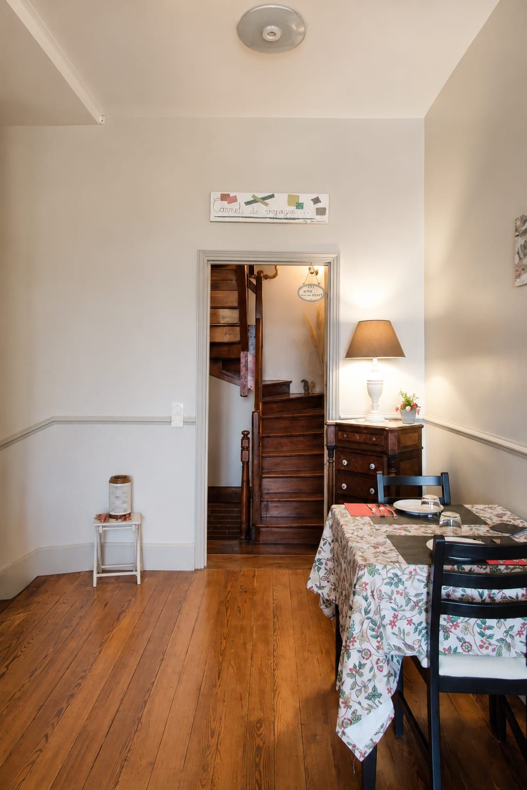 A dining area with a table set for two, a wooden staircase, and a small white table with a basket.