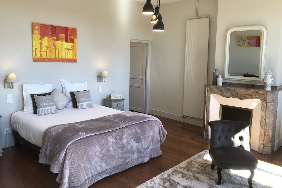 A bedroom with a bed, bedside lamps, a nightstand, a fireplace, a chair, and a painting on the wall.