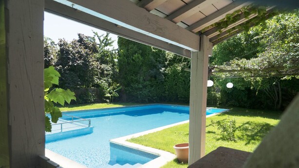 Una piscina rectangular con una escalera metálica, rodeada por un muelle de madera y una zona cubierta.