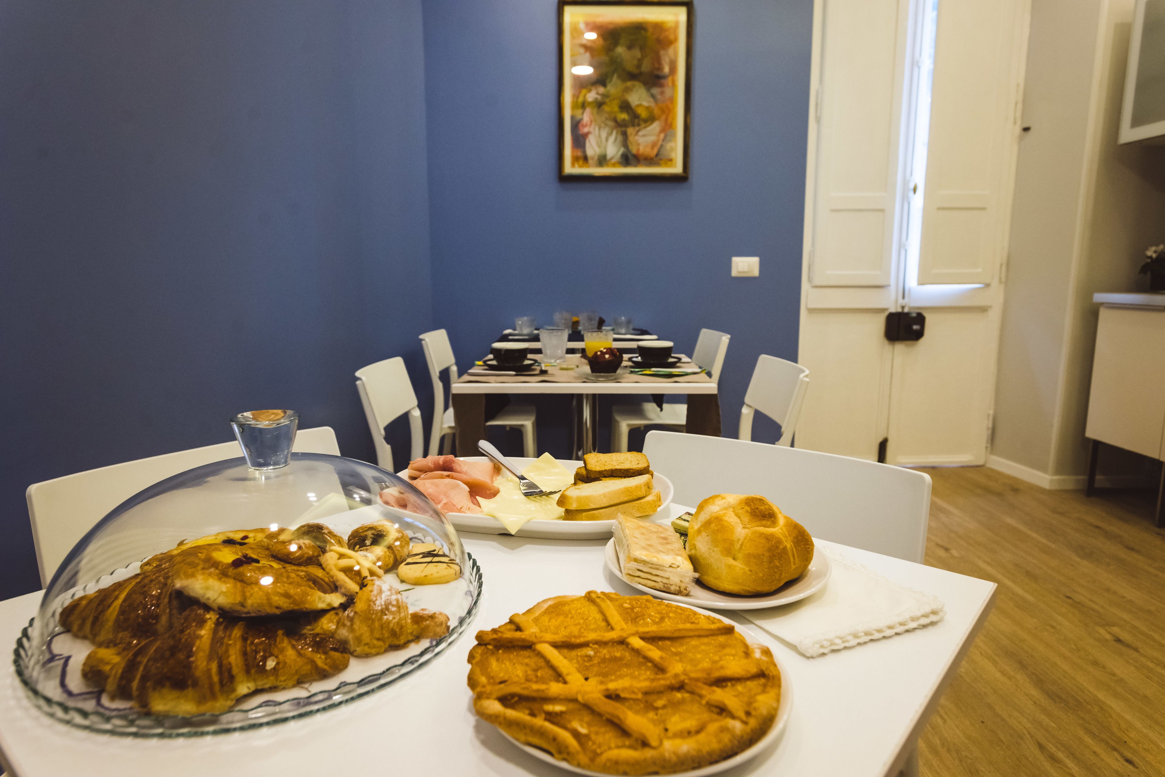 A dining table with a variety of food items, including a covered dish, bread, and a pie, in a room with blue walls and a painting on the wall.