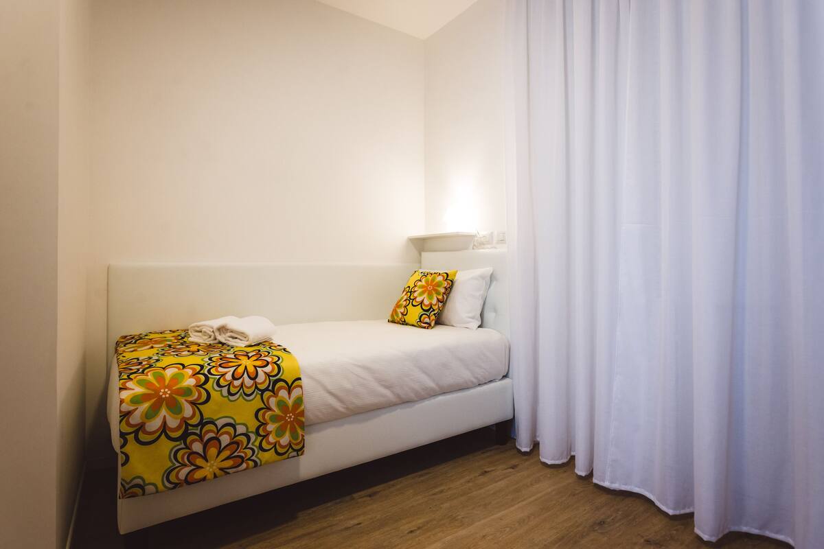 A single bed with a floral bedspread, white pillows, and a bedside table with a lamp.