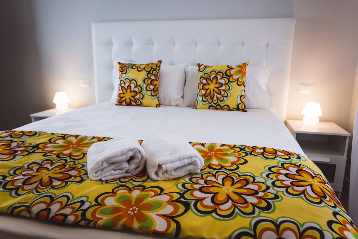 A neatly made bed with a floral bedspread, two white pillows, and a folded towel on the bed.