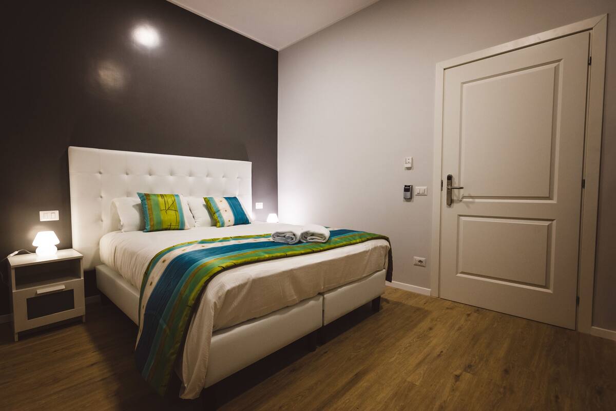 A bedroom with a white bed, a nightstand with a lamp, and a door.