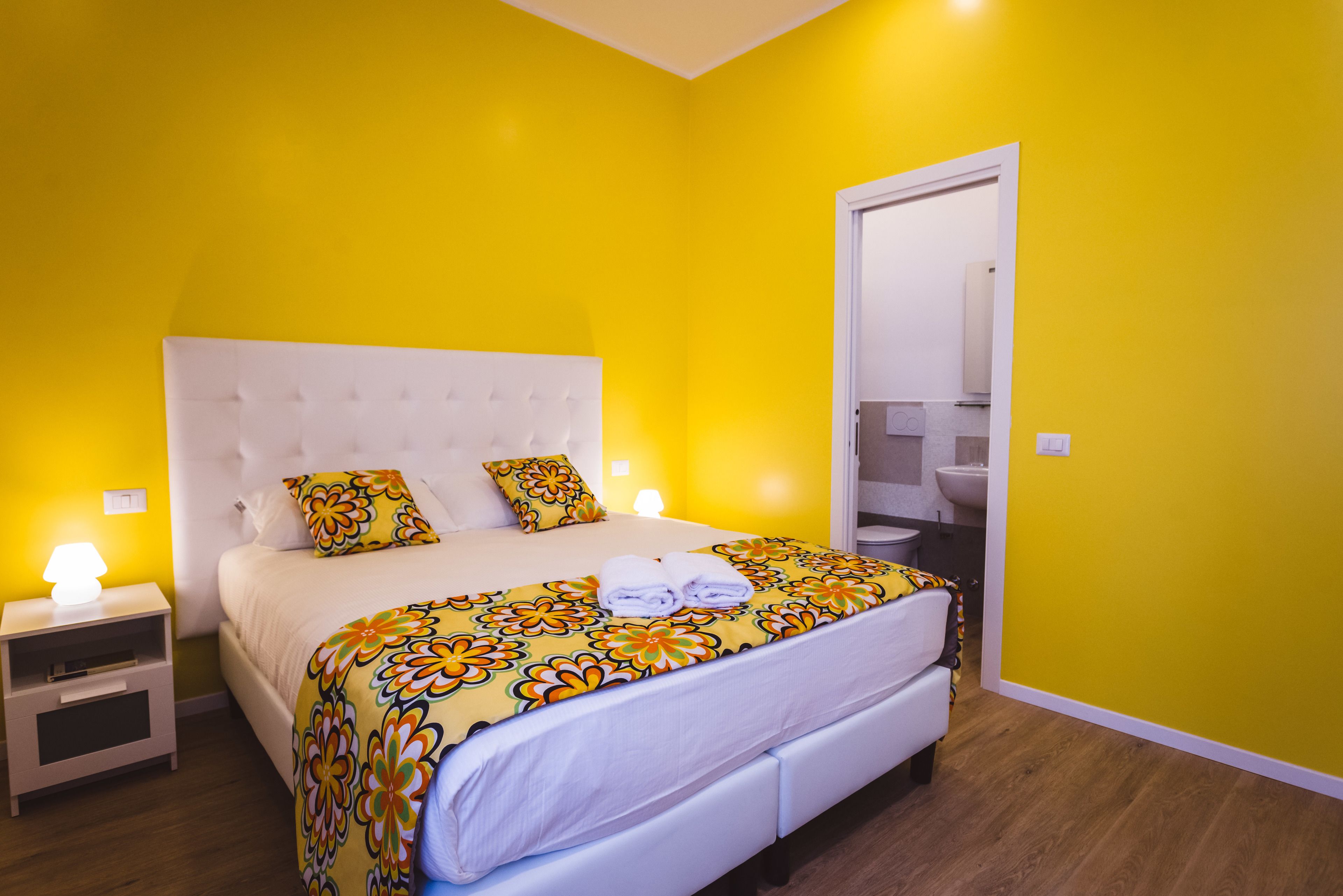 Een heldergele kamer met een groot bed, een wit hoofdeinde en een nachtkastje met een lamp.
