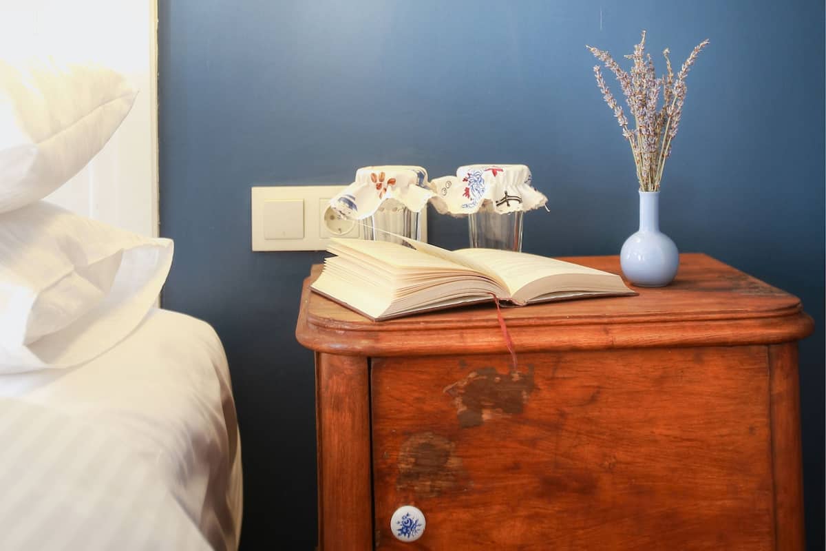 A bedroom with a bedside table, a lamp, a vase with dried flowers, and a bed.