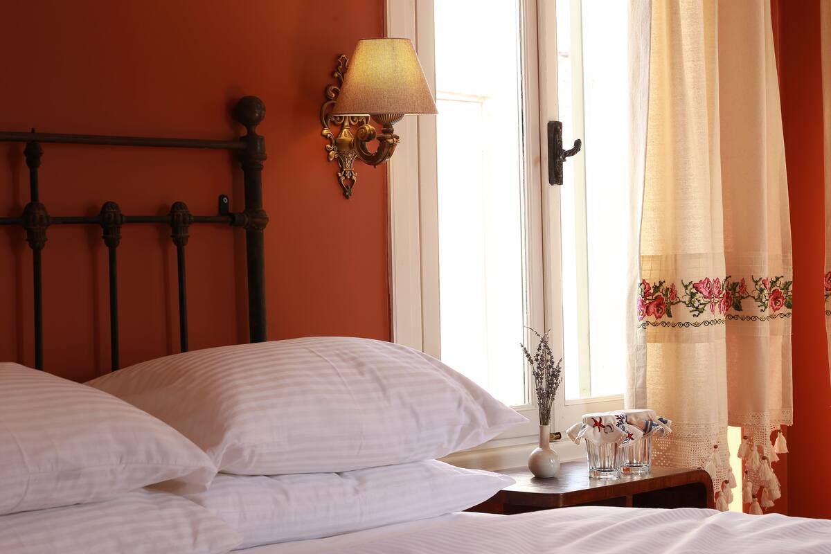 A neatly made bed with white linens, a wall-mounted lamp, and a window with sheer curtains.